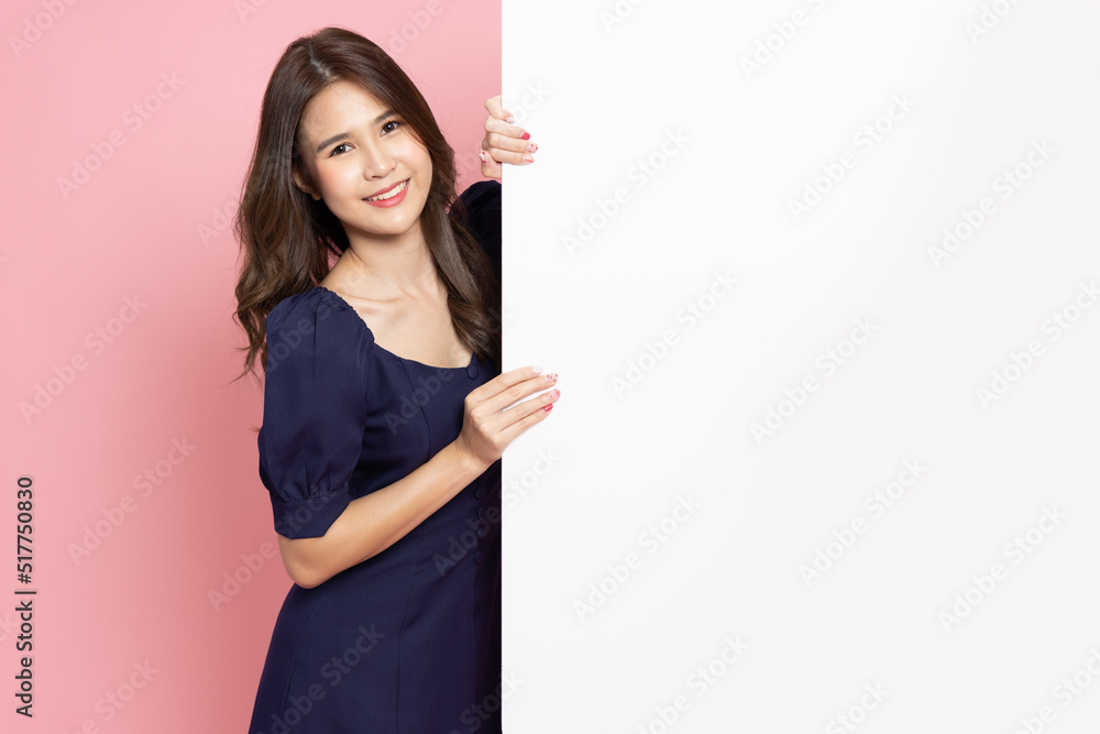 Happy young Asian woman is standing behind the white blank banner or empty copy space advertisement board on pink background, Looking at camera Happy young Asian woman is standing behind the white blank banner or empty copy space advertisement board on pink background, Looking at camera