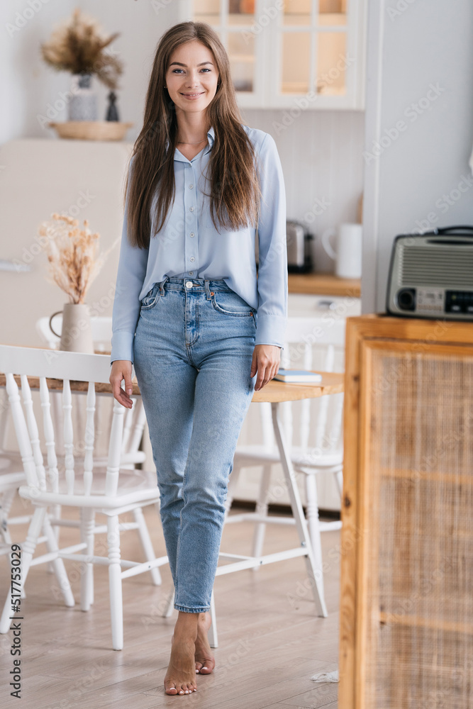 Vertical shot of beautiful girls in jeans and a blue shirt standing in the kitchen, looking at the camera, smiling contentedly. Beautiful satisfied european blonde housewife home relaxing on weekend. Vertical shot of beautiful girls in jeans and a blue shirt standing in the kitchen, looking at the camera, smiling contentedly. Beautiful satisfied european blonde housewife home relaxing on weekend.