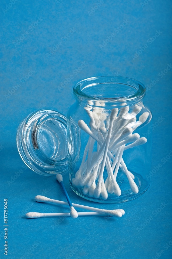 Open glass apothecary jar filled with cotton swabs with adjoining lid and three swabs on blue background