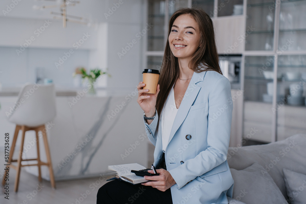 Beautiful Swedish girl with long hair in blue jacket, black trousers, sitting o sofa, holding coffee against blurry of kitchen with daily planner, cup of coffee, looking at the camera. Happy female. Beautiful Swedish girl with long hair in blue jacket, black trousers, sitting o sofa, holding coffee against blurry of kitchen with daily planner, cup of coffee, looking at the camera. Happy female.