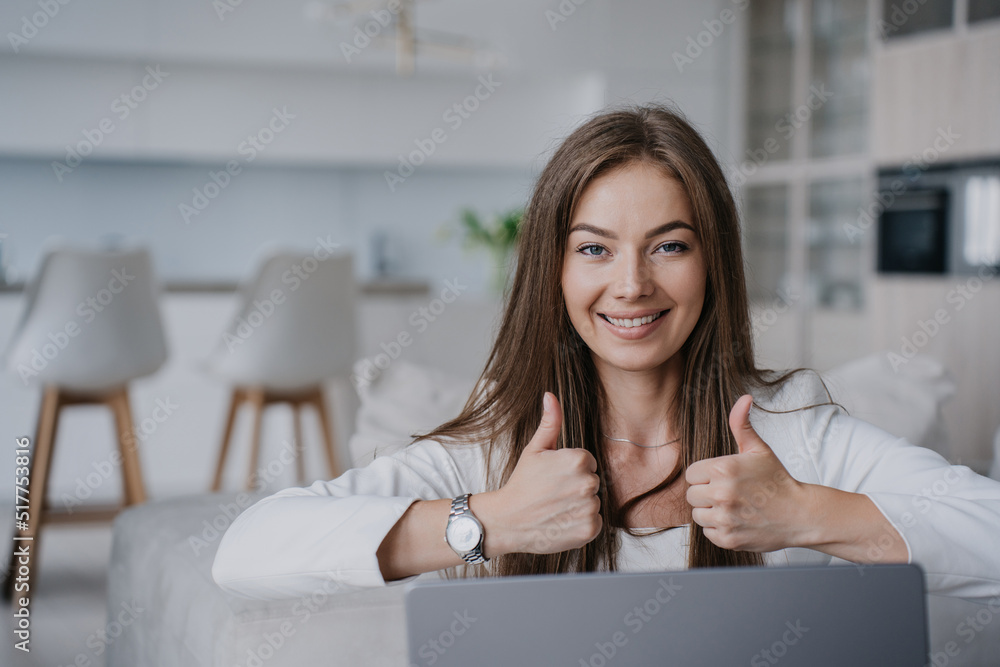 Beautiful blonde girl sitting at laptop, looking at the camera, showing thumbs up with both hands against background of a blurry kitchen at home. Cheerful European student has finished her studies. Beautiful blonde girl sitting at laptop, looking at the camera, showing thumbs up with both hands against background of a blurry kitchen at home. Cheerful European student has finished her studies.