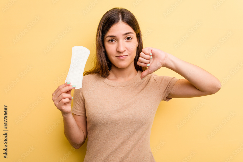 Young caucasian woman holding a sanitary napkin isolated on yellow background showing a dislike gesture, thumbs down. Disagreement concept. Young caucasian woman holding a sanitary napkin isolated on yellow background showing a dislike gesture, thumbs down. Disagreement concept.