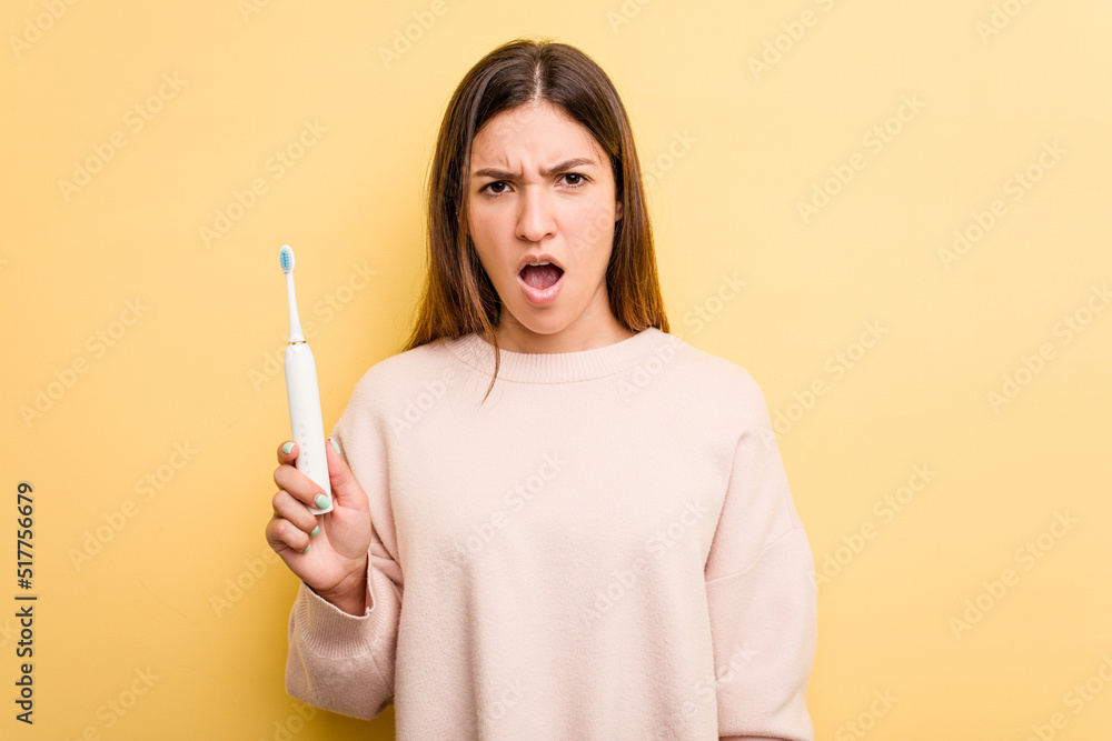 Young caucasian woman holding a electric toothbrush isolated on yellow background screaming very angry and aggressive. Young caucasian woman holding a electric toothbrush isolated on yellow background screaming very angry and aggressive.