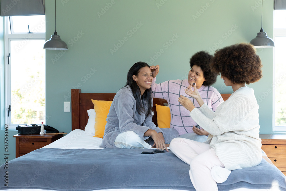 Biracial female friends applying make-up and painting fingernails of happy young woman on bed Biracial female friends applying make-up and painting fingernails of happy young woman on bed