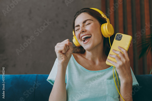 Young woman she 20s wearing casual clothes mint t-shirt headphones hold mobile cell phone sing song in microphone sit on blue sofa indoor rest at home in own room apartment. People lifestyle concept. photo