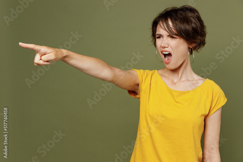 Young sad indignant angry mad furious woman she 20s wear yellow t-shirt point index finger aside scream shout isolated on plain olive green khaki background studio portrait. People lifestyle concept.