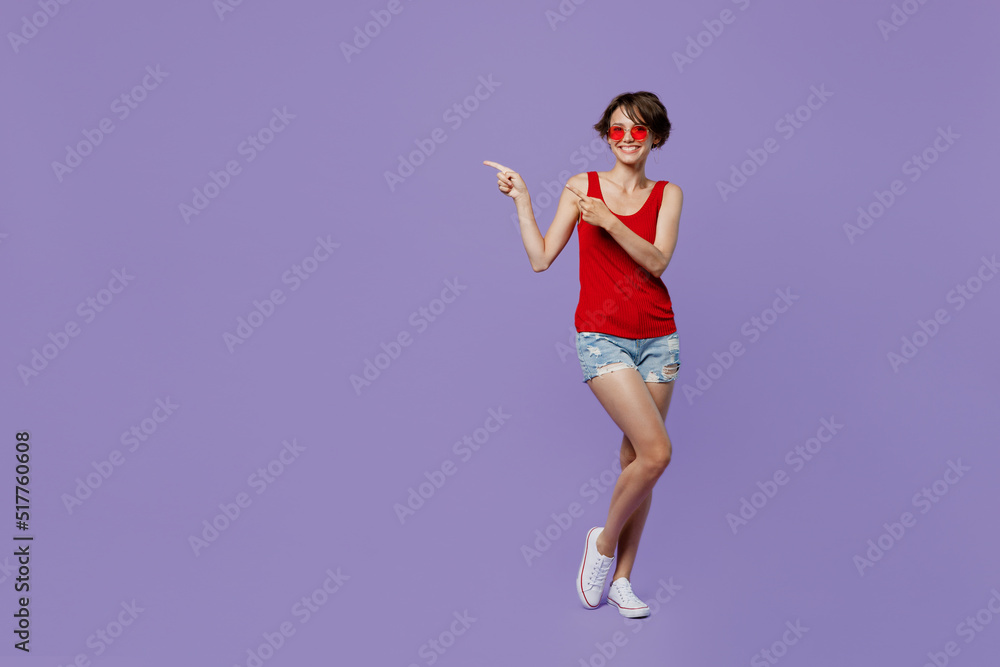 Full body young smiling fun woman 20s she wear red tank shirt eyeglasses pointing index finger aside indicate on workspace area copy space mock up isolated on plain purple backround studio portrait