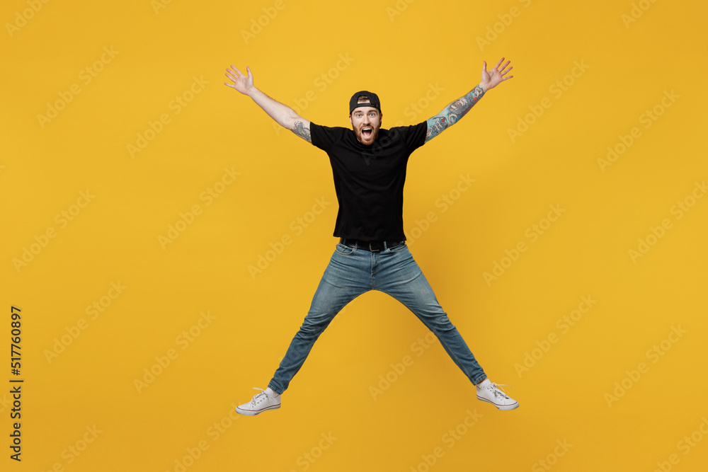 Full body young overjoyed excited exultant happy jubilant bearded tattooed man 20s he wears casual black t-shirt cap jump high with outstretched hands legs isolated on plain yellow wall background