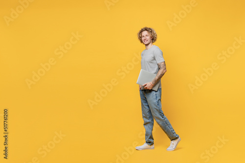 Full body young student smiling cheerful fun caucasian man 20s he wear grey t-shirt look camera hold use closed laptop pc computer isolated on plain yellow backround studio. People lifestyle concept.