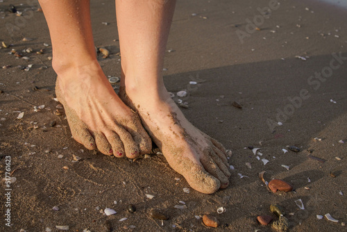 Detail of Two Barefoot Female Feet with Painted Nails Perched on the Sand with Shells and Pebbles on the Beach. Concept of Taking Care of Shoes in Summer