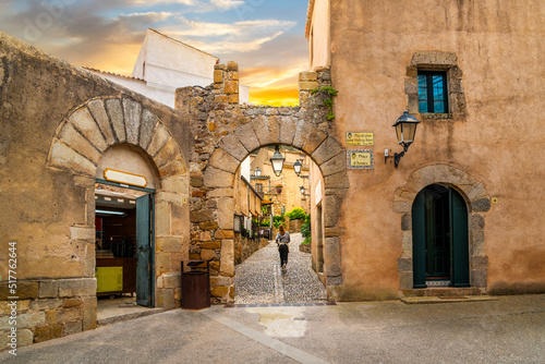 Fotografie, Obraz A woman walks through an arched passage on a cobblestone alley in the historic medieval hilltop village of Tossa de Mar on the Costa Brava coast of Spain, at sunset