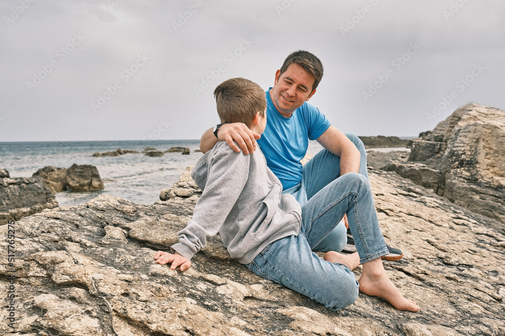 Happy family, father and son bonding, sitting on stone by the sea looking at view enjoying summer vacation. Togetherness Friendly concept Happy family, father and son bonding, sitting on stone by the sea looking at view enjoying summer vacation. Togetherness Friendly concept