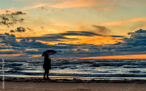 Colorful sunset on sandy beach of the Baltic Sea in Jurmala - famous tourist resort in Latvia, selective focus on silhouette of woman with umbrella