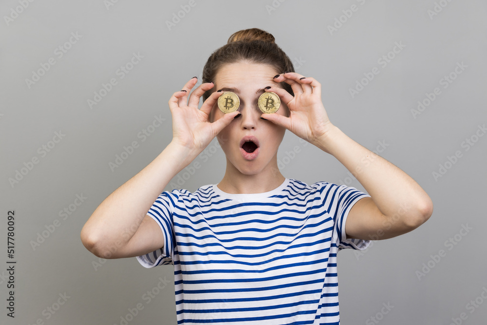 Portrait of amazed surprised woman wearing striped T-shirt with bun hairstyle covering eyes with golden bitcoin, having shocked expression. Indoor studio shot isolated on gray background. Portrait of amazed surprised woman wearing striped T-shirt with bun hairstyle covering eyes with golden bitcoin, having shocked expression. Indoor studio shot isolated on gray background.