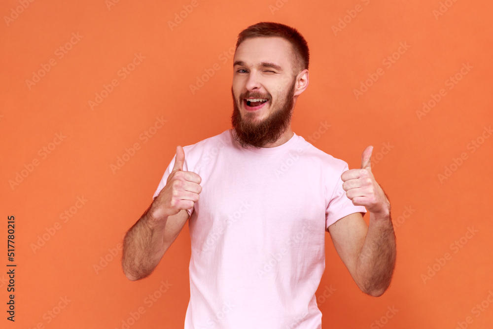 Portrait of positive bearded man standing with thumbs up, like gesture, demonstrating approval and agree with suggestion, wearing pink T-shirt. Indoor studio shot isolated on orange background. Portrait of positive bearded man standing with thumbs up, like gesture, demonstrating approval and agree with suggestion, wearing pink T-shirt. Indoor studio shot isolated on orange background.