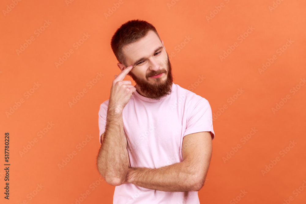 Portrait of bearded man contemplating leaning head on hand, pondering difficult question, trying to make right choice, wearing pink T-shirt. Indoor studio shot isolated on orange background. Portrait of bearded man contemplating leaning head on hand, pondering difficult question, trying to make right choice, wearing pink T-shirt. Indoor studio shot isolated on orange background.