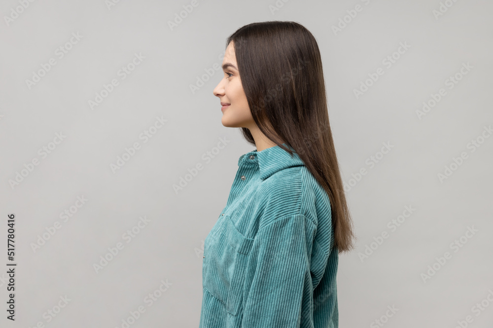 Side view of happy successful young woman standing looking at camera with charming and joyous toothy smile, wearing casual style jacket. Indoor studio shot isolated on gray background. Side view of happy successful young woman standing looking at camera with charming and joyous toothy smile, wearing casual style jacket. Indoor studio shot isolated on gray background.