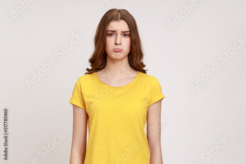 Portrait of sad beautiful teenager girl wearing yellow T-shirt looking at camera with pout lips and upset facial expression, being offended. Indoor studio shot isolated on gray background.