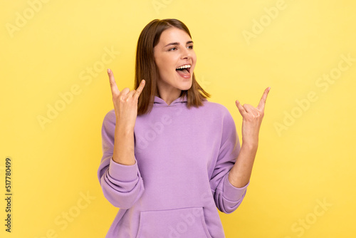 Excited enthusiastic woman with dark hair showing rock and roll gesture. screaming rapturously, looking away, wearing purple hoodie. Indoor studio shot isolated on yellow background.