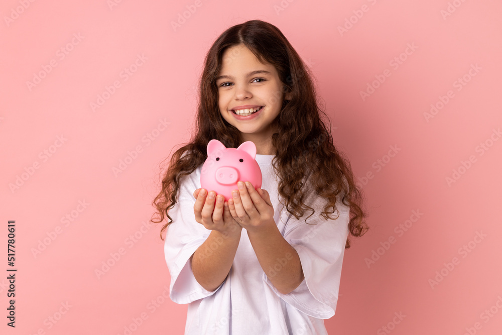Portrait of little girl wearing white T-shirt holding pink piggy bank, looking at camera with charming smile, best variant for saving money. Indoor studio shot isolated on pink background. Portrait of little girl wearing white T-shirt holding pink piggy bank, looking at camera with charming smile, best variant for saving money. Indoor studio shot isolated on pink background.
