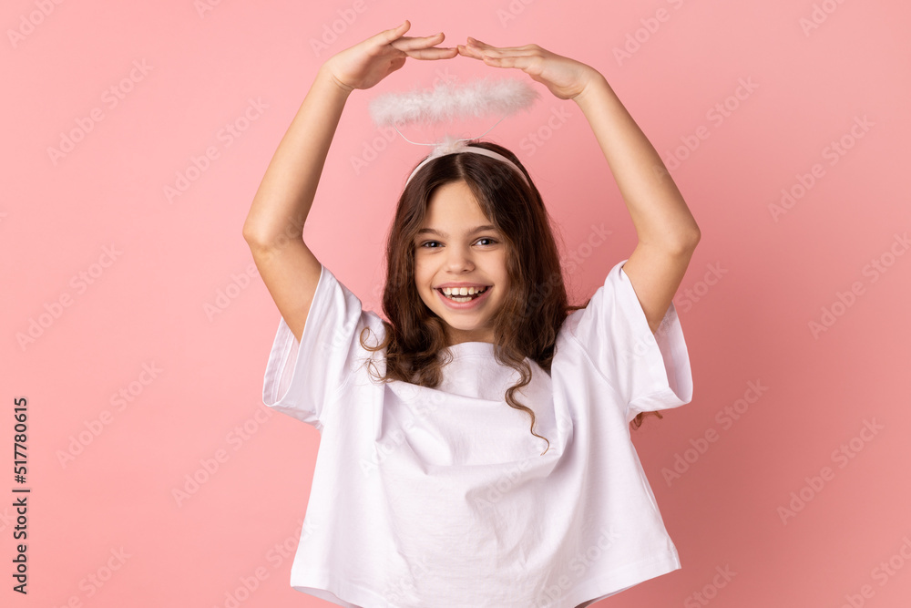 Portrait of little girl wearing white T-shirt and halo standing with raised arms, looking at camera with toothy smile, expressing positive emotions. Indoor studio shot isolated on pink background. Portrait of little girl wearing white T-shirt and halo standing with raised arms, looking at camera with toothy smile, expressing positive emotions. Indoor studio shot isolated on pink background.