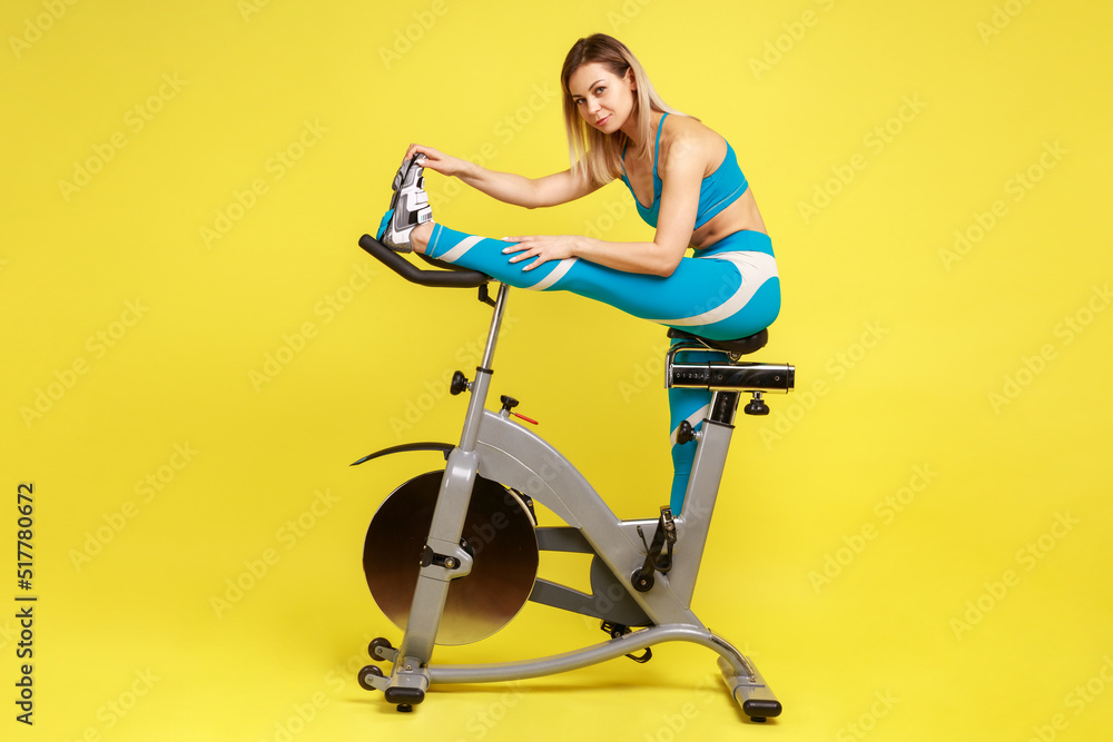 Side view portrait of woman with blonde hair sitting on exercise bike and stretching leg, warming up before cardio workout, wearing blue sportswear. Indoor studio shot isolated on yellow background. Side view portrait of woman with blonde hair sitting on exercise bike and stretching leg, warming up before cardio workout, wearing blue sportswear. Indoor studio shot isolated on yellow background.