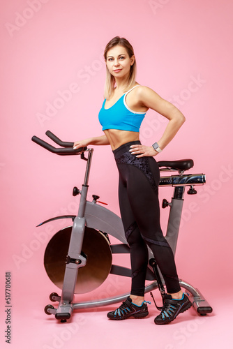 Full length portrait of charming woman with slim body shape posing near exercise bike and looking at camera  wearing sports tights and top. Indoor studio shot isolated on pink background.