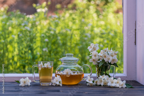 Hot herbal tea in glass teapot, cup and beautiful bouquet of jasmine flowers on windowsill at summer day near garden