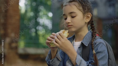 A little girl closeup standing on the schoolyard outside looking at food and eating a sandwich with chease, ham and salad. Food and nutrition concept. photo