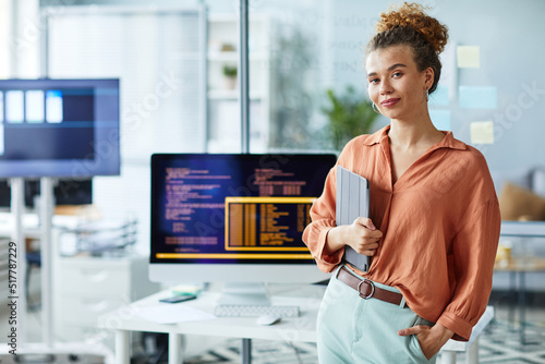 Portrait of young female programmer with digital tablet looking at camera while standing at her workplace with computer