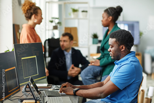 African young programmer writing codes on computer sitting at table with colleagues talking in background