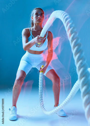 Beautiful sporty young woman with ponytail exercises with rope. Girl is dressed in white sports uniform and stands on blue background.