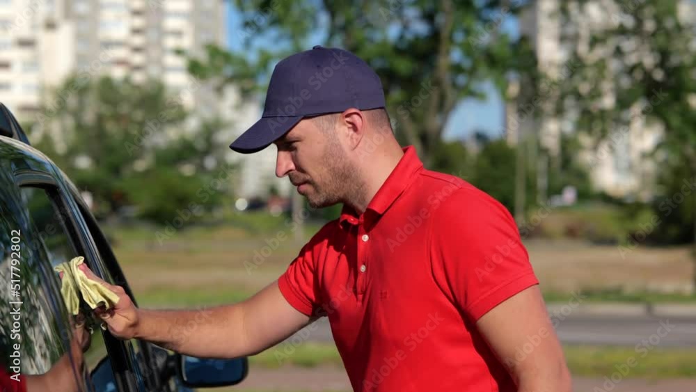 Close up of happy Caucasian handsome man wiping the glass and the body of the car standing on street. Male worker cleaning car outside. Side view. Maintenance. Work concept Close up of happy Caucasian handsome man wiping the glass and the body of the car standing on street. Male worker cleaning car outside. Side view. Maintenance. Work concept