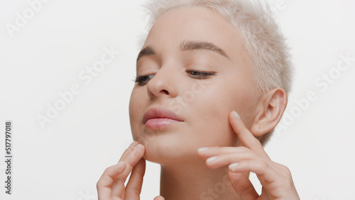Big close-up shot of gorgeous young slim Caucasian woman with short light hair strokes her face on white background | Skin hydration concept