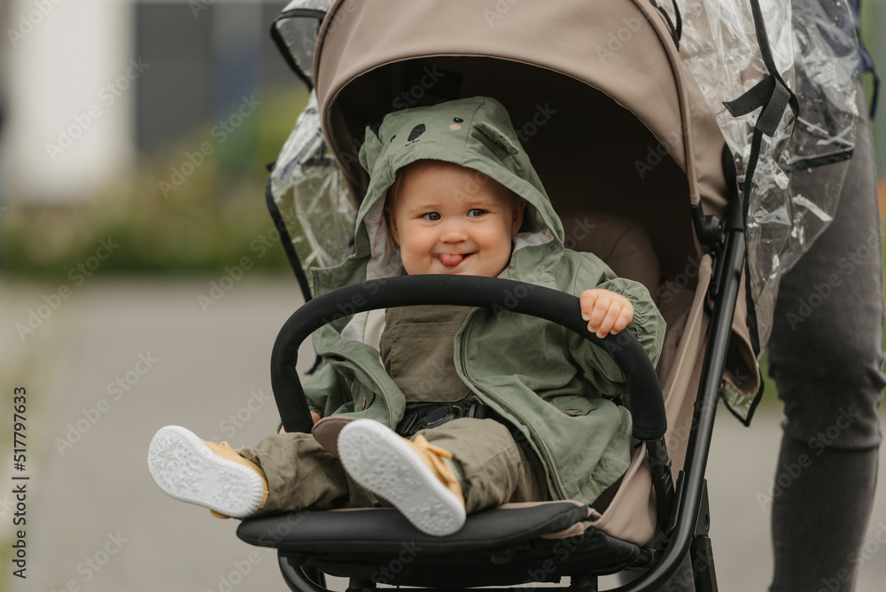 A female toddler with her tongue out is sitting in the stroller on a cloudy day. A young girl in a baby carriage in a village green.