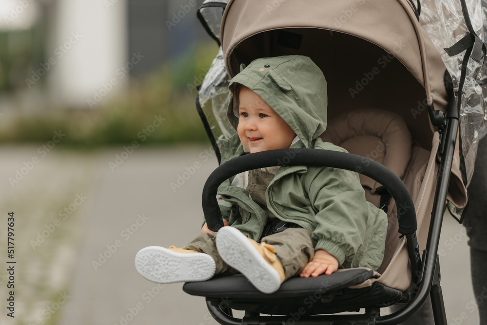 A happy female toddler is sitting in the stroller on a cloudy day. In a green village, a young girl in a raincoat is relaxing in a baby carriage.