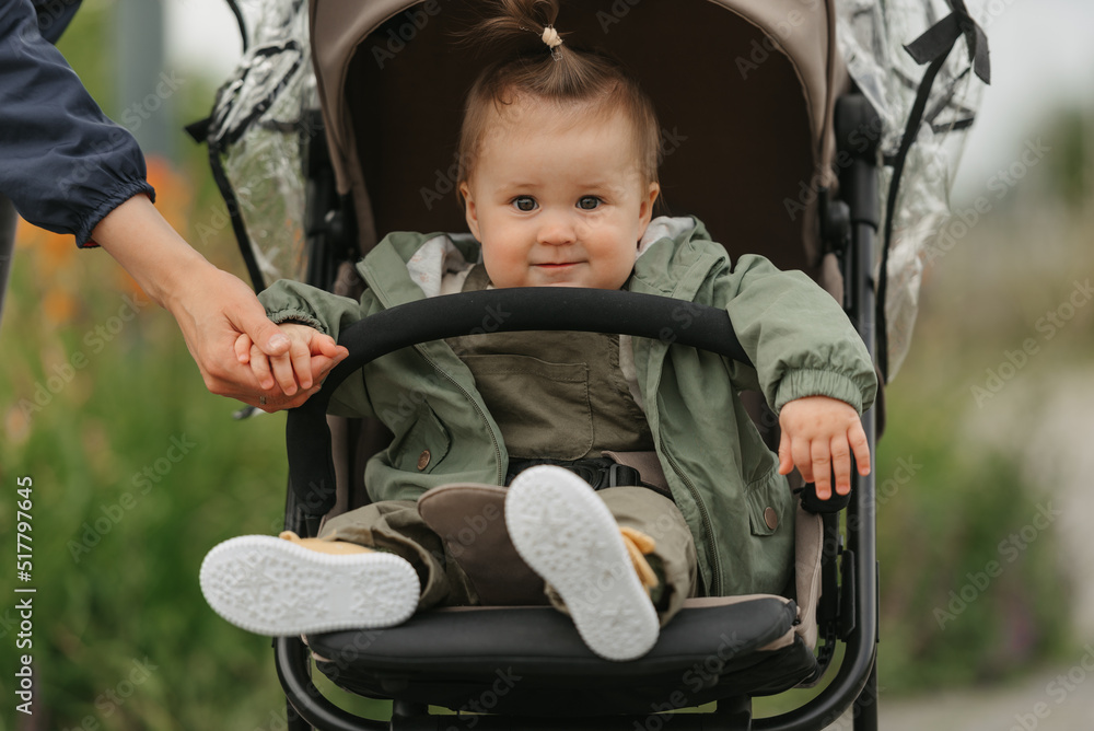 A female toddler is sitting in the stroller on a cloudy day. In a green village, a young pretty girl in a raincoat is in a baby carriage. A mother is holding the hand of her daughter. A female toddler is sitting in the stroller on a cloudy day. In a green village, a young pretty girl in a raincoat is in a baby carriage. A mother is holding the hand of her daughter.