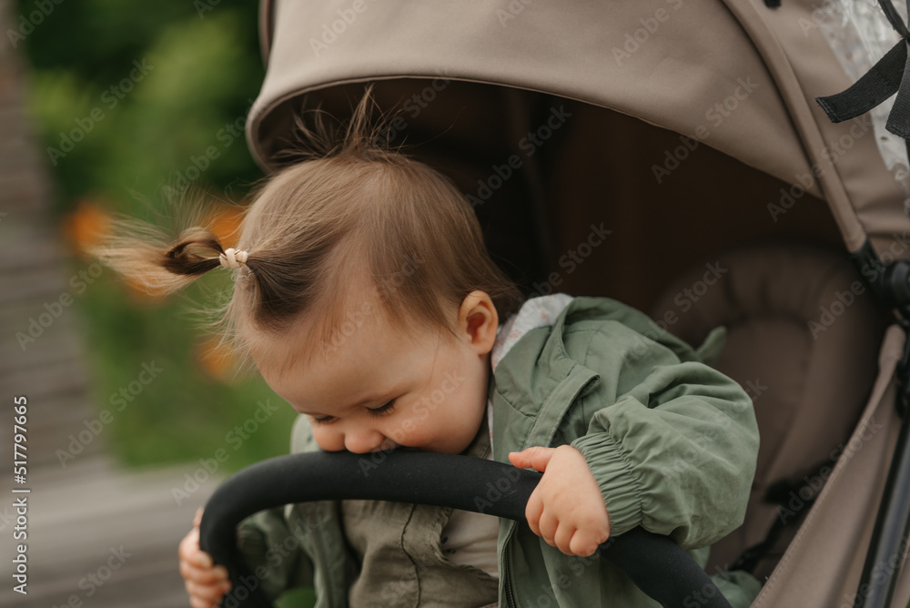 A close portrait of a female toddler who is eating the safety bumper bar of the stroller on a cloudy day. A young girl in a raincoat is in her baby carriage in a park at noon. A close portrait of a female toddler who is eating the safety bumper bar of the stroller on a cloudy day. A young girl in a raincoat is in her baby carriage in a park at noon.