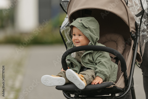 A happy female toddler is sitting in the stroller on a cloudy day. In a green village, a young girl in a raincoat is relaxing in a baby carriage.