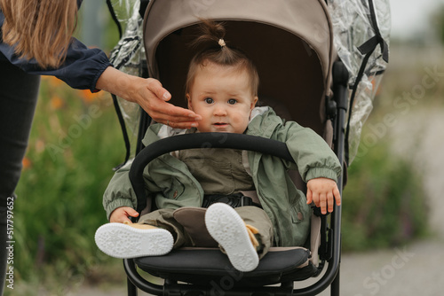A female toddler is sitting in the stroller on a cloudy day. In a green village, a young serious girl in a raincoat is in a baby carriage. A mother is touching the cheek of her daughter.
