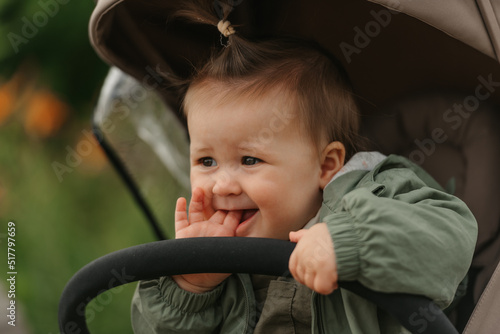 A close portrait of a female toddler who is putting her fingers in her mouth in the stroller. A young girl in a raincoat with open eyes is in her baby carriage in a park at noon.
