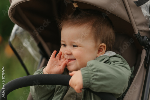 A close portrait of a thoughtful female toddler who is putting her fingers in her mouth in the stroller on a cloudy day. In a green village, a young girl in a raincoat is in her baby carriage.