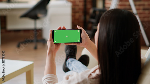 Woman sitting on sofa while holding smartphone with green screen mockup template background. Young adult person with modern touchscreen mobile phone having chroma key isolated display.