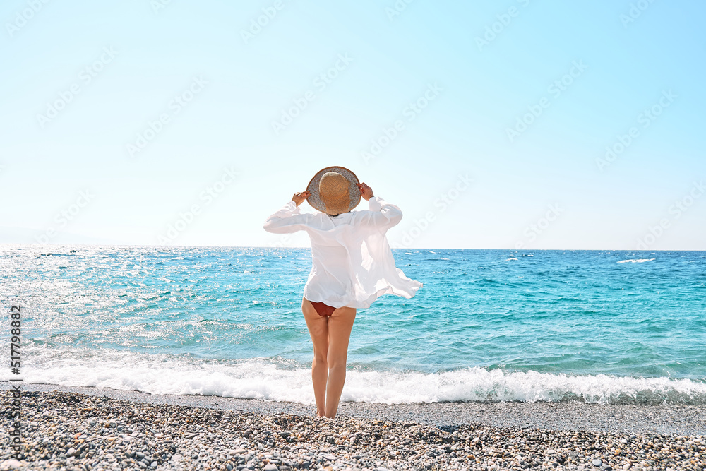 Back view of carefree woman in straw hat and white shirt walking on pebble beach. Relaxing and enjoy holiday at the sea in summertime. Unity with nature. Wellness, success, freedom and travel concept Back view of carefree woman in straw hat and white shirt walking on pebble beach. Relaxing and enjoy holiday at the sea in summertime. Unity with nature. Wellness, success, freedom and travel concept