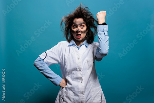 Crazy scientist with dirty face and wacky hair angrily clenching and raising fist in air while gritting teeth. Funny looking mad infuriated biochemist looking at camera on blue background. Studio shot