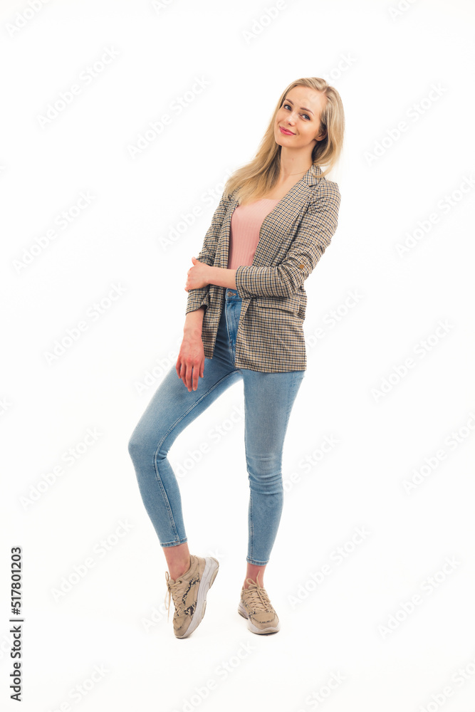 Cute caucasian blond woman in her 20s wearing jeans, sneakers, and checked blazer, standing, posing, and looking at camera with a smile. Full body isolated copy space studio shot over white background Cute caucasian blond woman in her 20s wearing jeans, sneakers, and checked blazer, standing, posing, and looking at camera with a smile. Full body isolated copy space studio shot over white background