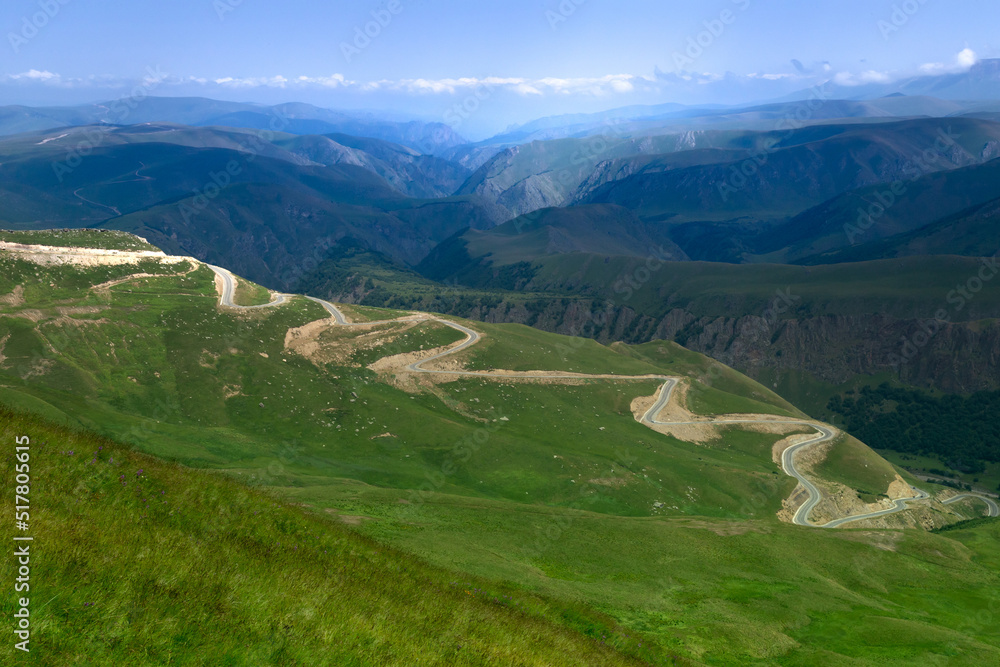 Naklejka premium winding mountain road with serpentines, green valley, high blue mountains, majestic nature, mountain landscape, travel, tourism, road adventure, direction, space for text, wallpaper, summer