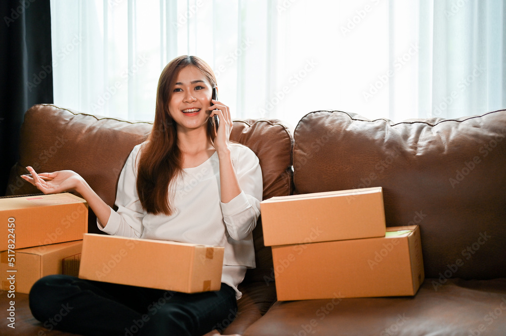 Attractive young asian woman sitting on sofa with cardboard boxes and talking on the phone Attractive young asian woman sitting on sofa with cardboard boxes and talking on the phone