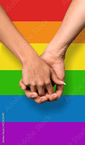 Two hands holding each other with the LGBTQ rainbow flag in the background. Symbol for homosexual community for pride month and the celebration day of sexual and love diversity