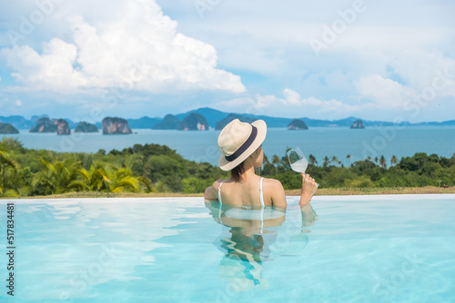 Happy woman in white swimsuit drinking wine in luxury swimming pool hotel against beautiful view, young female with hat enjoy in tropical resort. Relaxing, summer travel, holiday and vacation concept